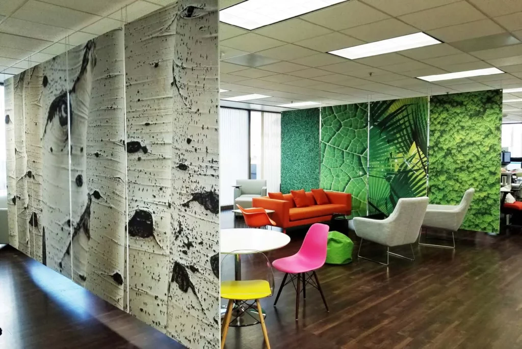 Modern office lounge with colorful chairs, orange sofas, and walls decorated with large nature-themed murals—including green leaves, moss, and birch bark patterns—creating an inviting setting reminiscent of premier suites.