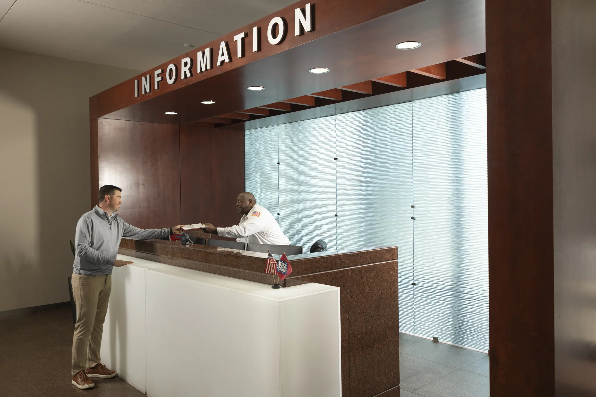 A man stands at an information desk, handing an item to an attendant behind the counter in a modern office lobby, where the Simmons Tower amenities add convenience and comfort to every visitor’s experience.