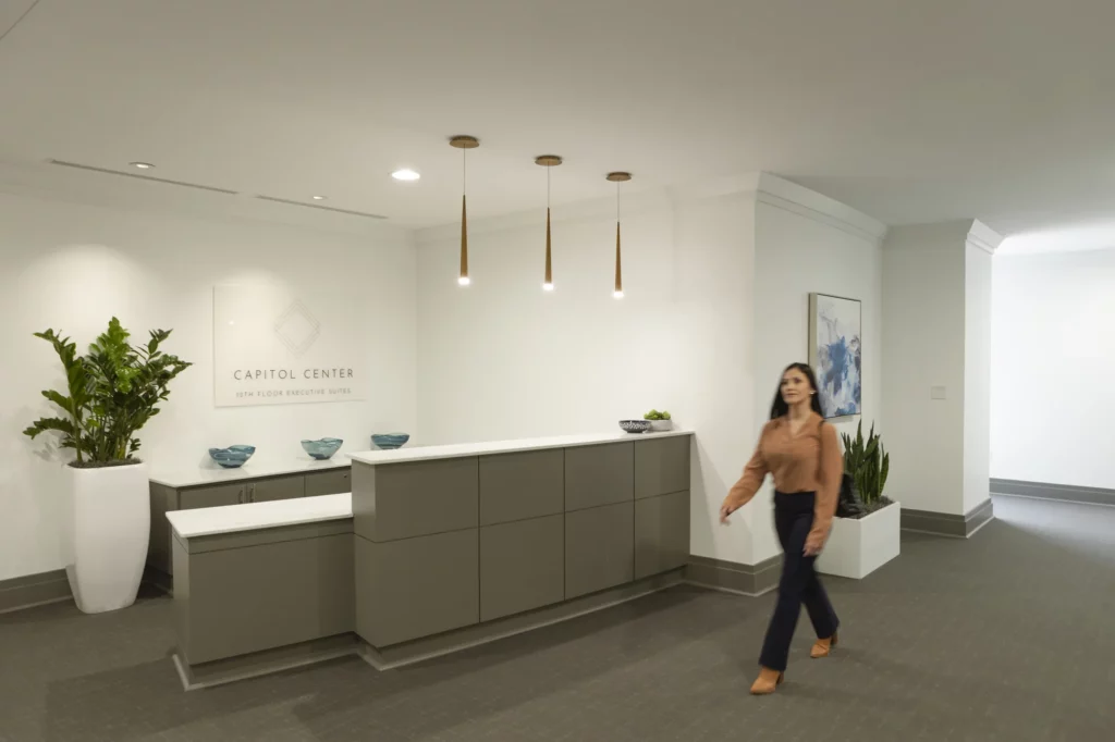 A woman walks past a modern executive suites reception area with a gray counter, green plants, and contemporary decor.