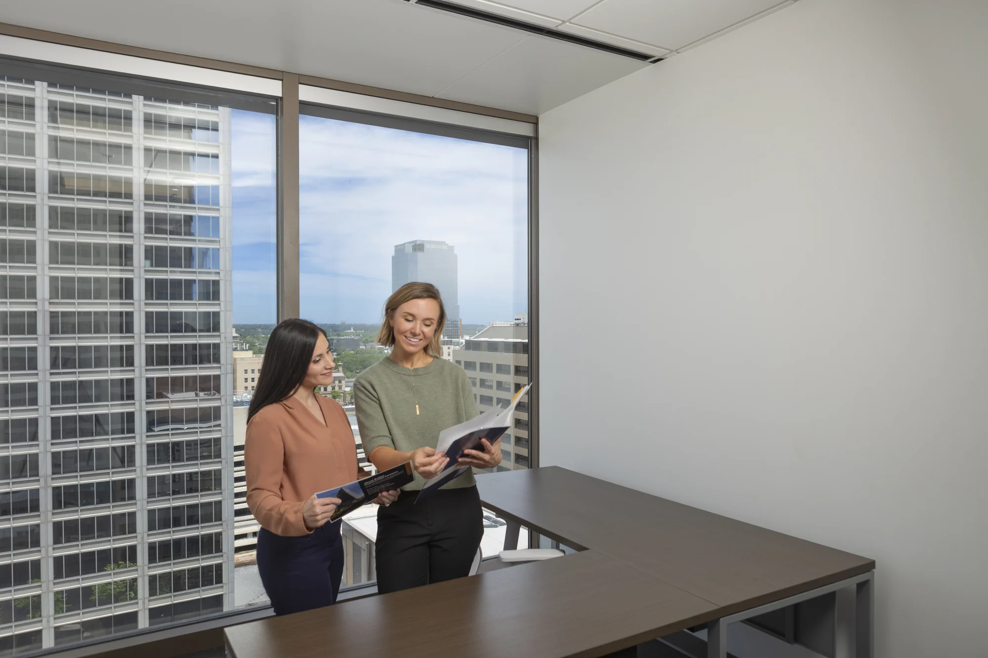 Two women stand in an office with large windows, reviewing documents together near a desk, with city buildings visible outside.