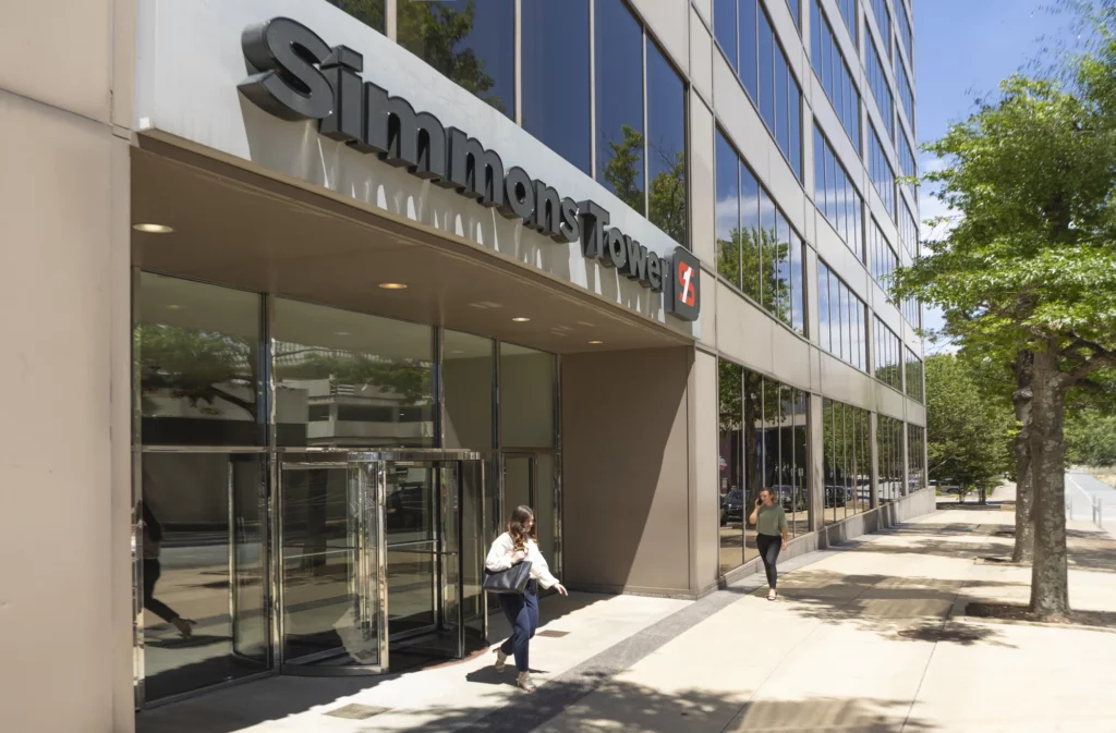 Two people walk outside the entrance of Simmons Tower, a tall office building with glass windows and a revolving door, on a sunny day.