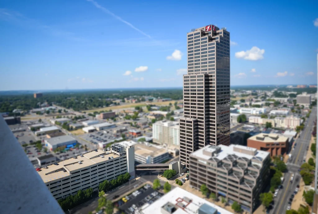 A tall office skyscraper, Simmons Tower, stands above nearby buildings on a sunny day, with a clear blue sky and city streets visible below.