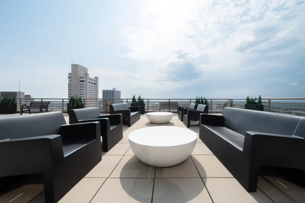 Rooftop patio with black benches, round white tables, potted plants, and a cityscape view under a partly cloudy sky, offering Simmons Tower amenities for an elevated urban experience.