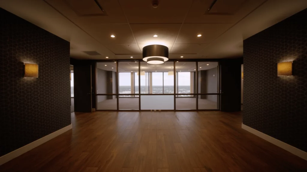 Empty office space with wood floors, dark walls, ceiling lights, a central hanging light fixture, and large glass windows overlooking the cityscape