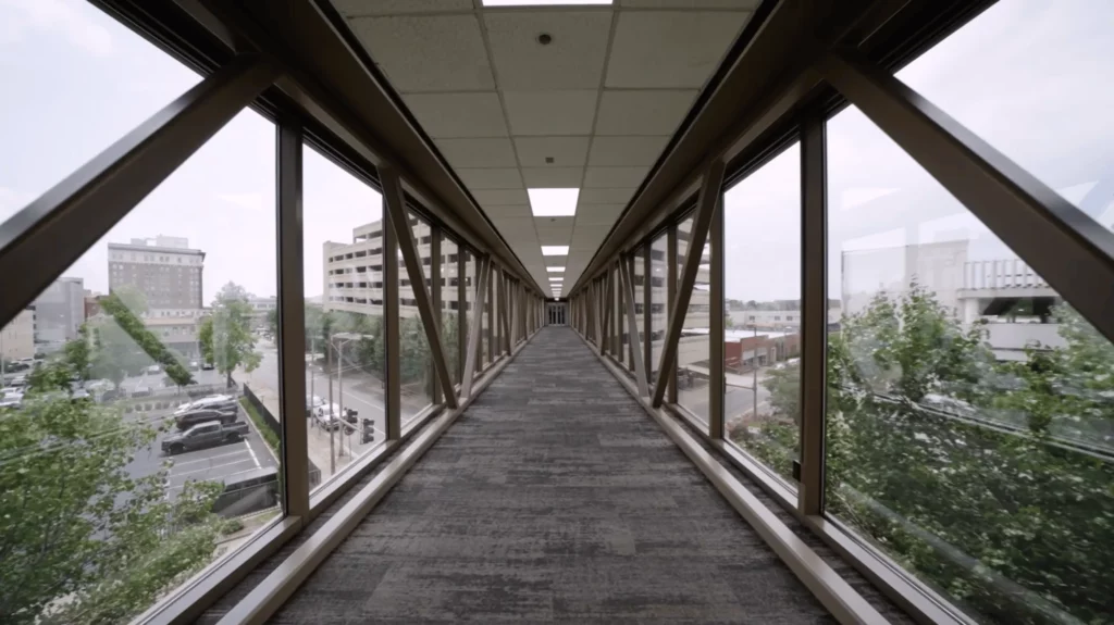 Enclosed pedestrian walkway with large windows on both sides, carpeted floor, and views of city buildings and streets outside.